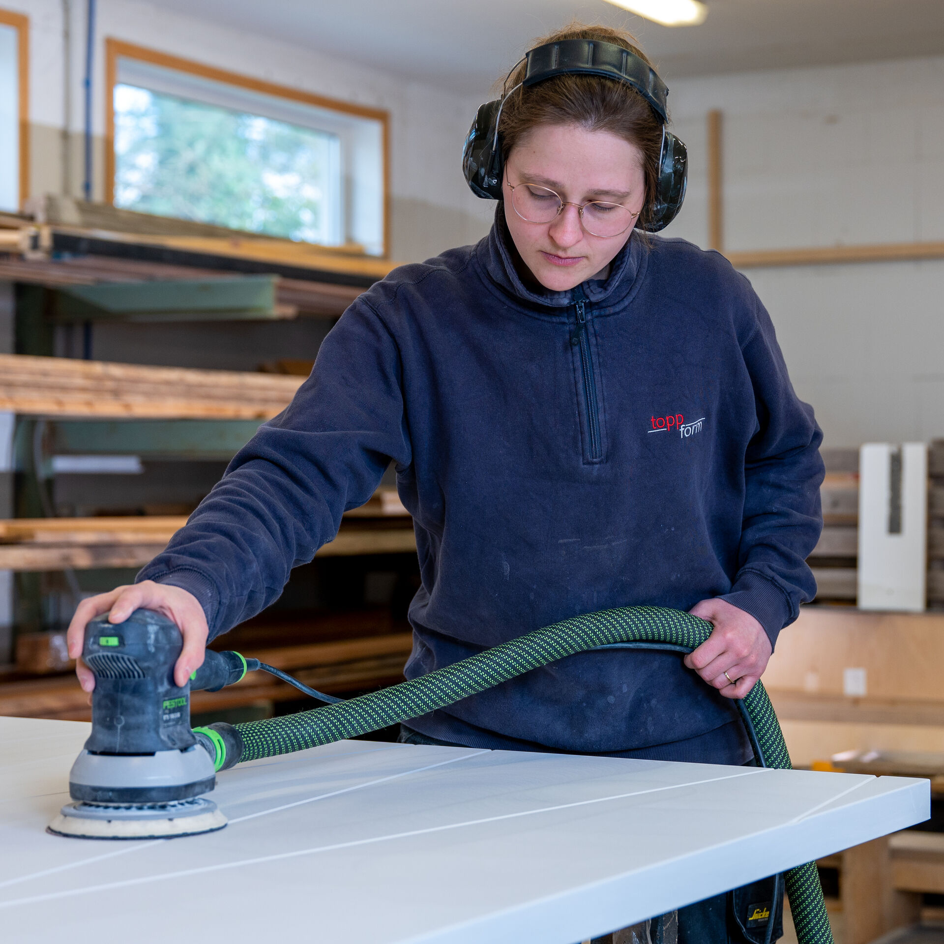 Rechts junge Frau, sie schleift Baumaterial, Hintergrund Regale mit Holzlatten